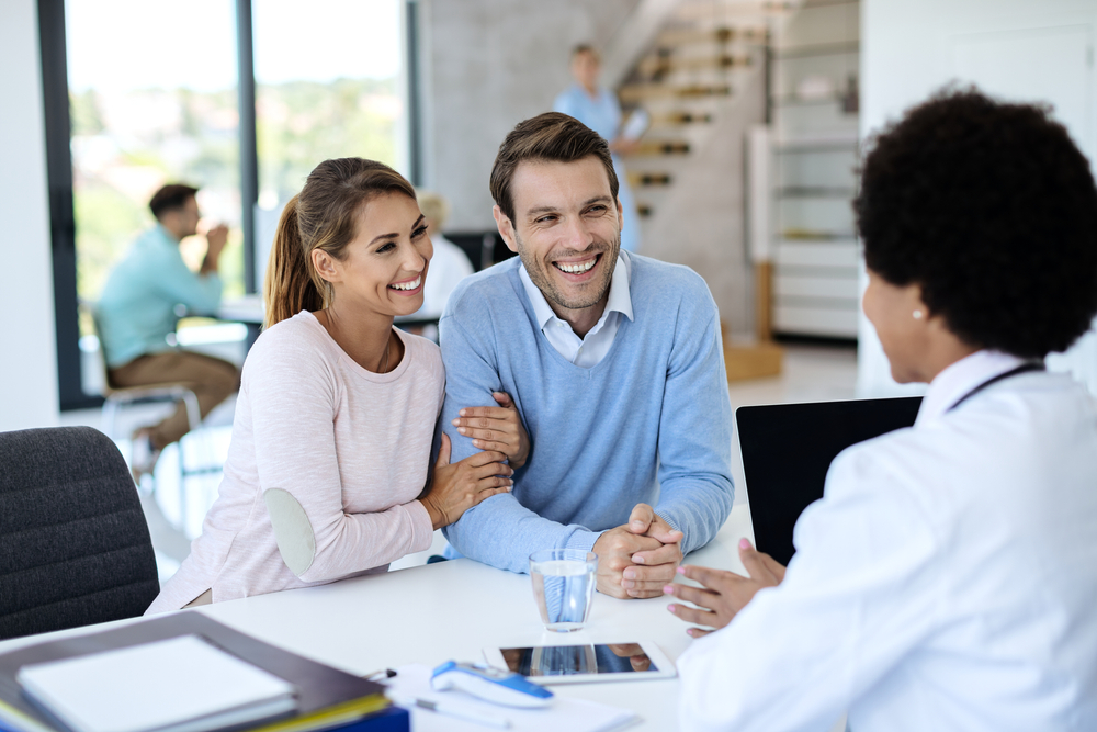 Young,Happy,Couple,Talking,To,Their,Doctor,While,Having,Appointment Coppia durante una visita preconcezionale con la ginecologa